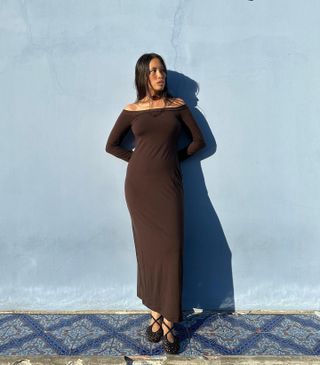 Fashion influencer Anne-Victoire Lefevre leaning against a baby blue wall wearing a stone pendant cord choker necklace, brown off-the-shoulder long-sleeve maxi dress, and black knit lace-up flats.