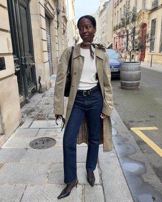 Fashion influencer Sylvie Mus poses on a Paris sidewalk wearing a tied silk neck scarf, a Burberry trench coat, ivory knit top, black shoulder bag, black belt, dark-wash straight-leg jeans, and black slingback heels.