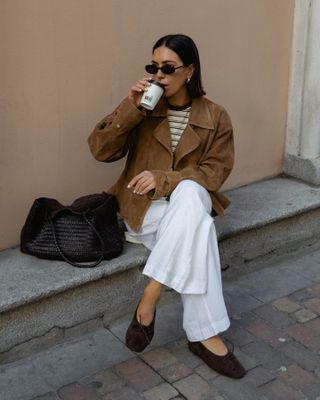 Fashion influencer Jessica Skye wearing oval sunglasses, a striped ringer tee, tan suede moto jacket, white linen pants, brown woven tote bag, and suede brown ballet flats.