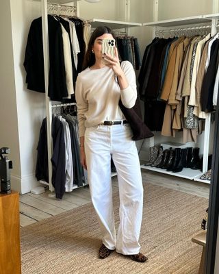 British fashion influencer Anna Newton poses for a mirror selfie with a wavy phone case, lightweight beige crewneck sweater, black belt with gold buckle and hardware, a brown suede tote bag, relaxed white jeans, and leopard-print flats.