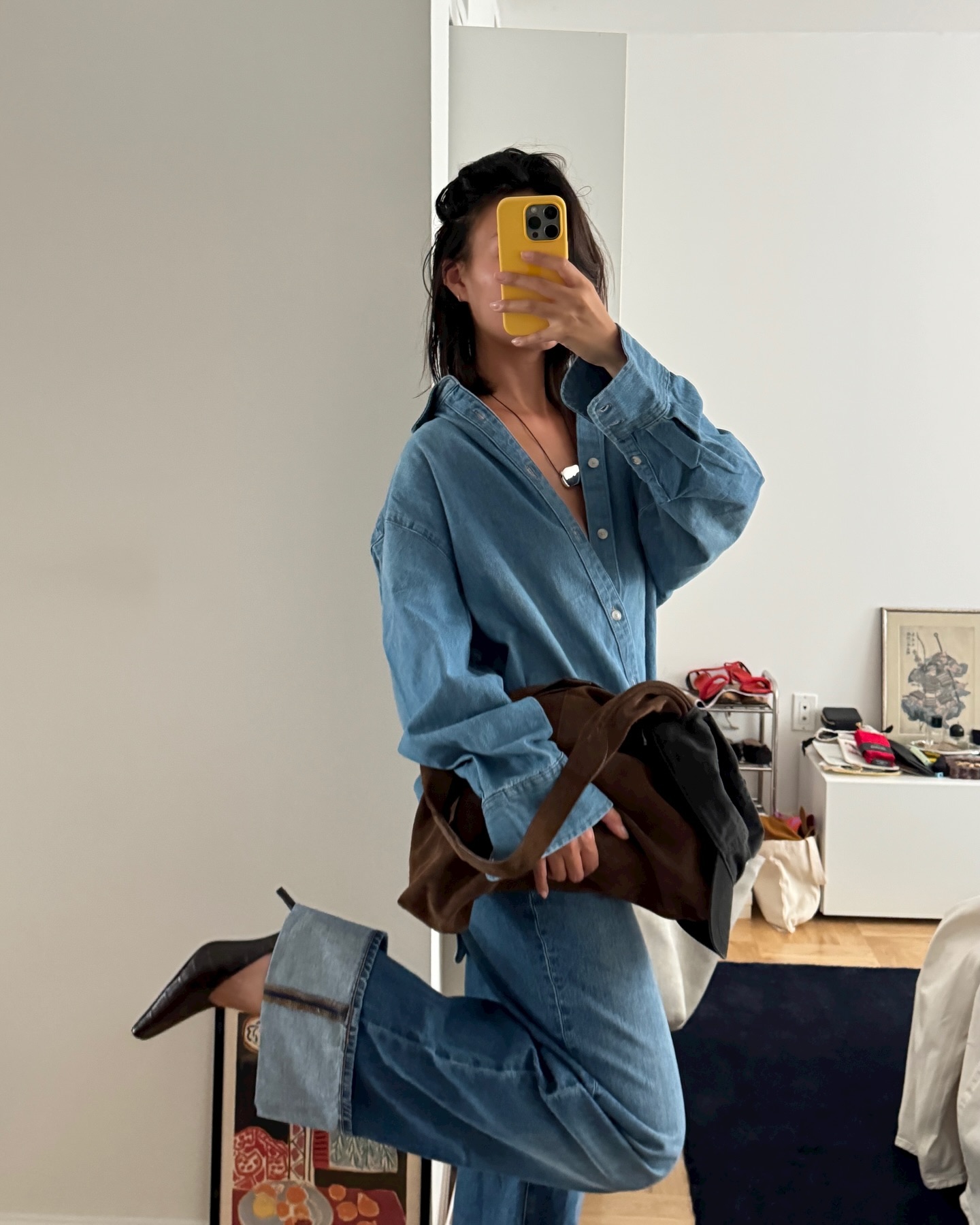style influencer Mimi Nguyen poses for a mirror selfie with a yellow phone case, a silver pendant cord necklace, denim button-down shirt, brown suede tote bag, wide-leg cuffed jeans, and black pointed-toe mule pumps