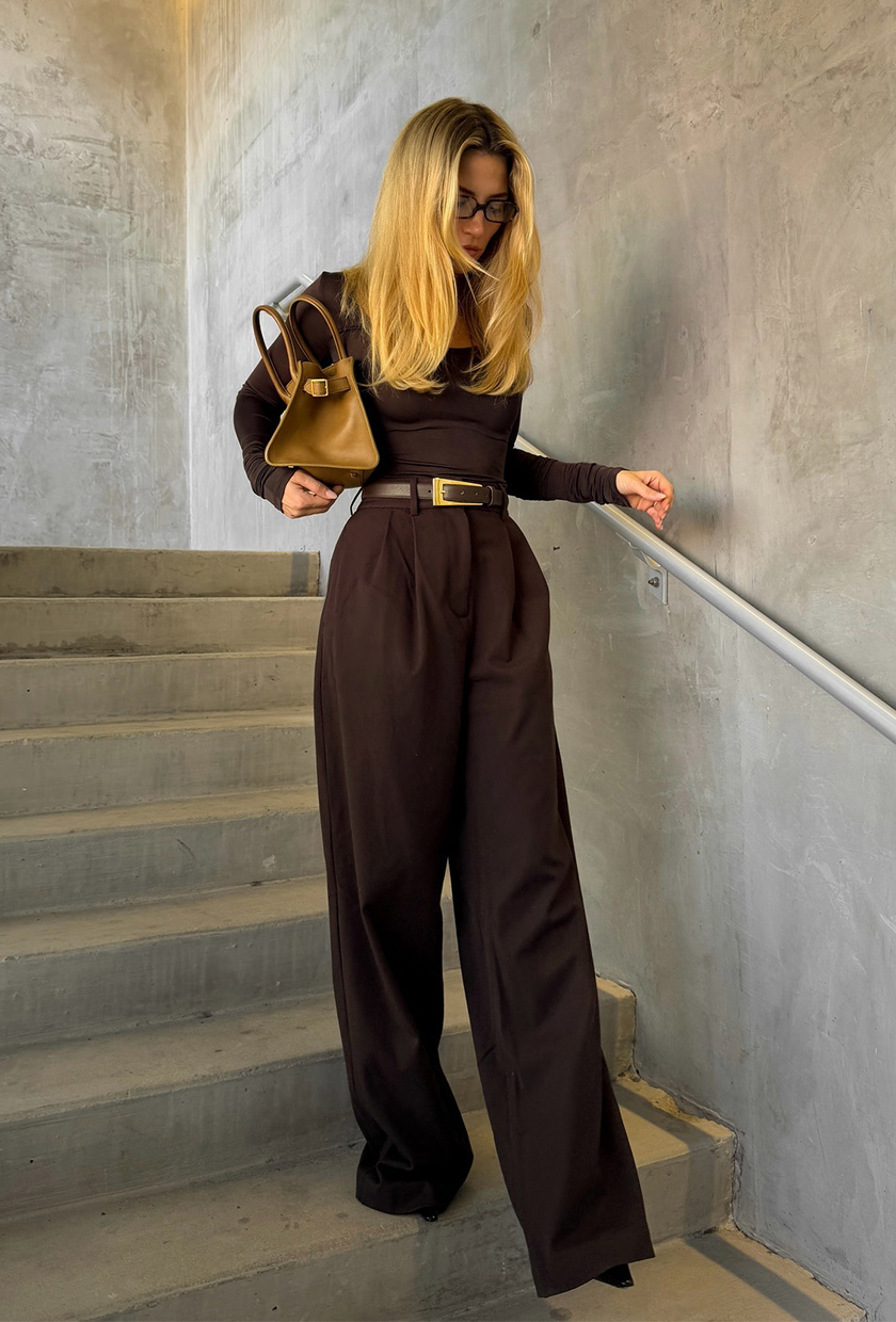 How to wear a statement belt is shown in a photo of a woman standing in a stairwell wearing a dark brown t-shirt with matching brown trousers, brown heels, a tan handbag, and a dark brown statement belt.