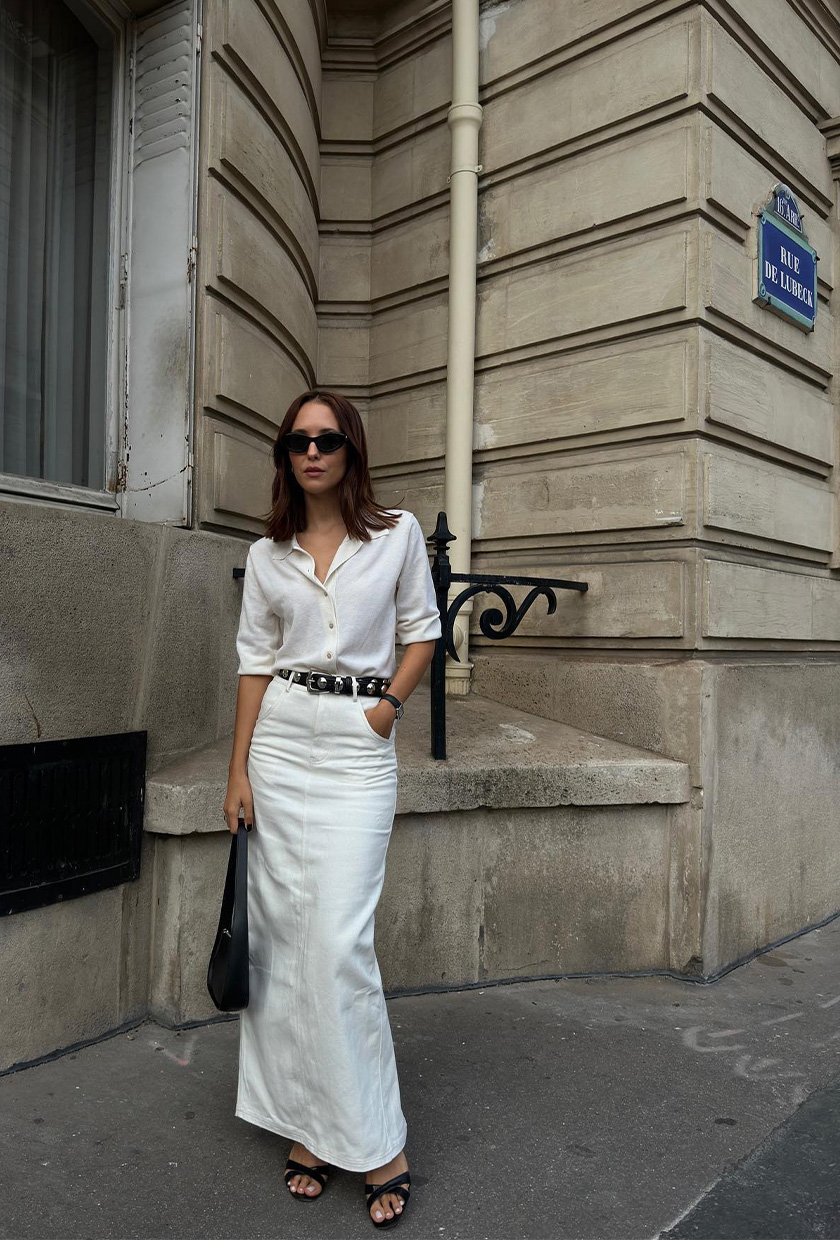 how to wear a statement belt is shown in a photo of a woman standing outside wearing a white button-down shirt, black studded belt, white maxi skirt, black heeled mules, and a black shoulder bag