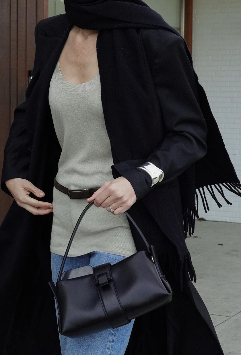 How to wear a statement belt is shown in a close photo of a woman standing outside wearing a black coat with a long scarf styled with a light gray tank top and jeans with a black bag, silver cuff bracelet, and a thin skinny statement belt