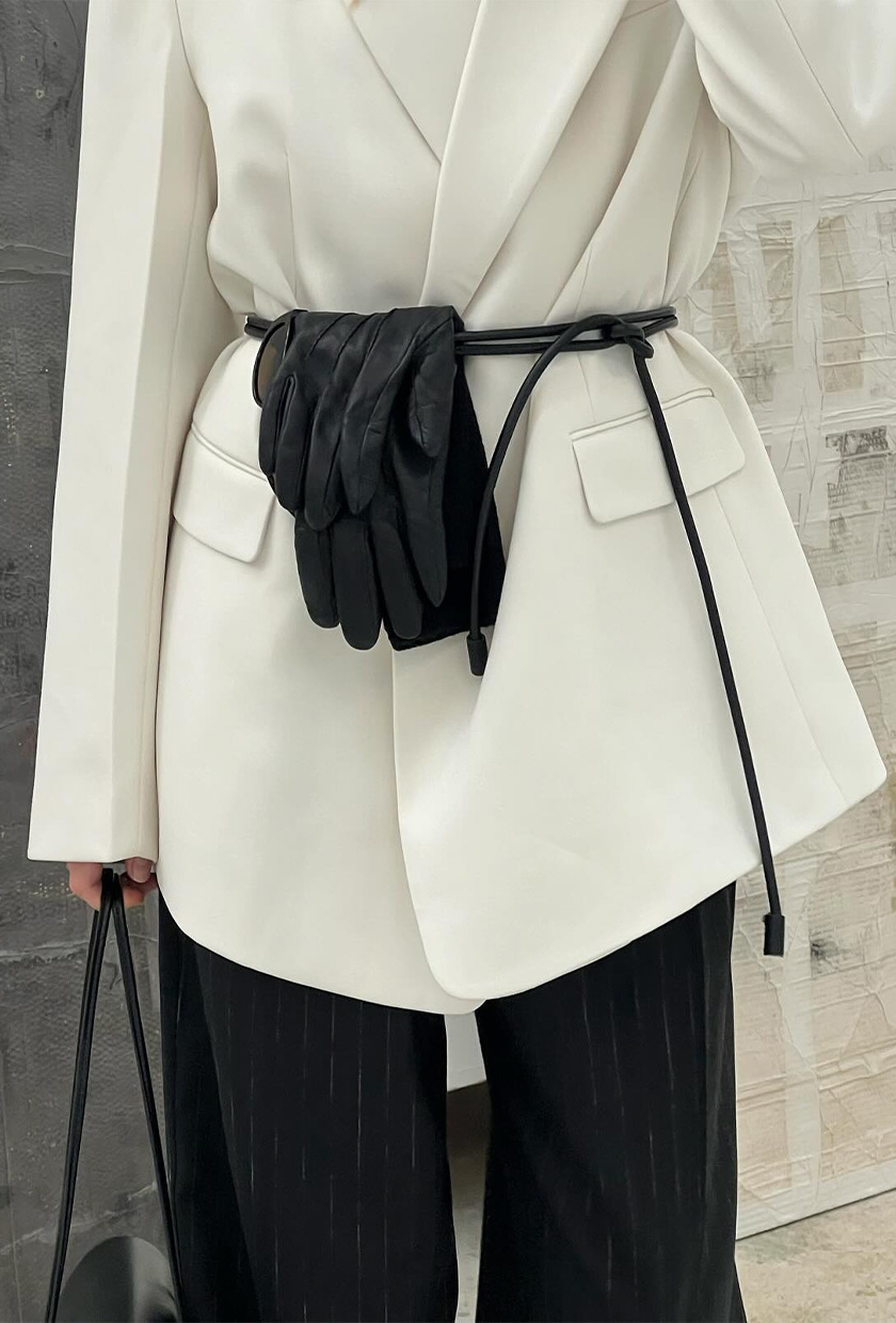 How to wear a statement belt is shown in a close photo of a woman wearing a white blazer with a thin rope belt tied on top with leather gloves tucked into the belt that's styled with black trousers and a black shoulder bag