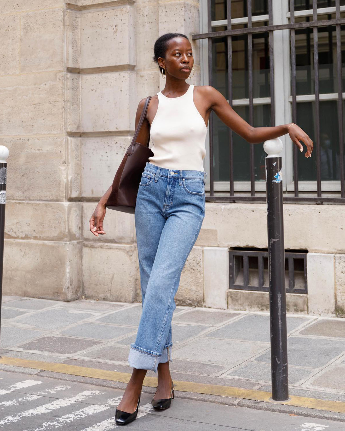style influencer Sylvie Mus wearing posing on a Paris sidewalk wearing a ivory racerback tank top, brown leather tote bag, cuffed straight-leg jeans, and black patent slingback kitten-heels.