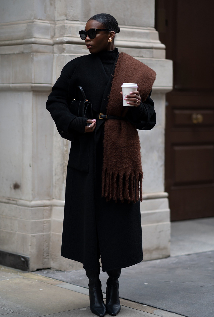 How to wear a statement belt is shown in a photo of a woman standing outside wearing a black sweater dress with a black long coat and black boots with a black statement belt layered on top with a brown chunky scarf tucked into the belt