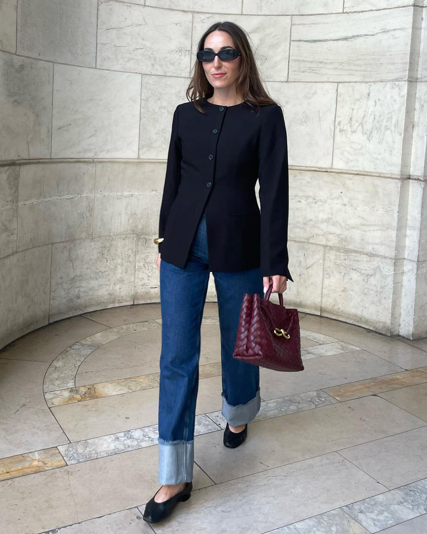 fashion editor Anna LaPlaca wearing black oval sunglasses, a crewneck cinched waist button-down blazer, burgundy Bottega Veneta bag, cuffed jeans, and black ballet flats
