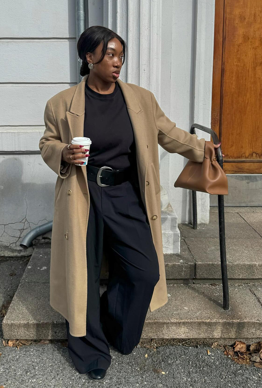 How to wear a statement belt is shown in a photo of a woman standing outside wearing a long tan coat over a black t-shirt with black trousers and black ballet flats with a tan handbag and a extra-wide black statement belt