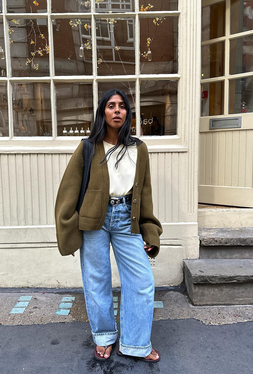 How to wear a statement belt is shown in a photo of a woman standing outside wearing a green chunky cardigan over a white silk high-neck tank top with high-rise cuffed jeans, brown flip-flops, a black tote back, and a black studded statement belt