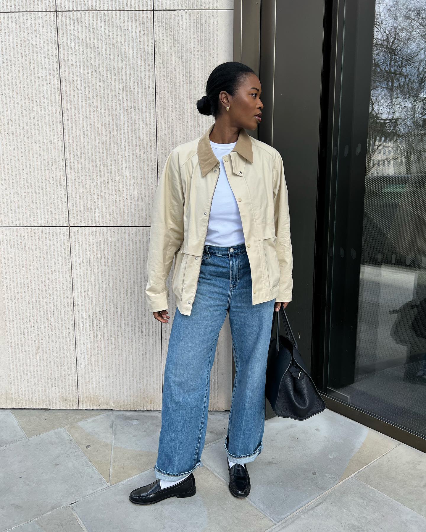 British fashion influencer Marilyn Nwawulor-Kazemaks poses wearing a beige barn jacket, white T-shirt, cuffed jeans, white socks, and black loafers