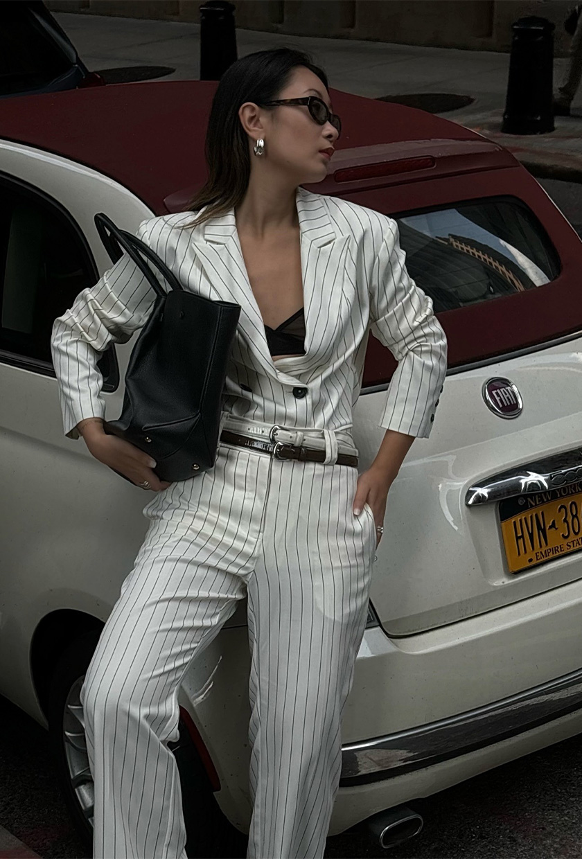 How to wear a statement belt is shown in a photo of a woman standing outside leaning against a Fiat car wearing a white pinstripe blazer with matching trousers and double-layered statement belts.