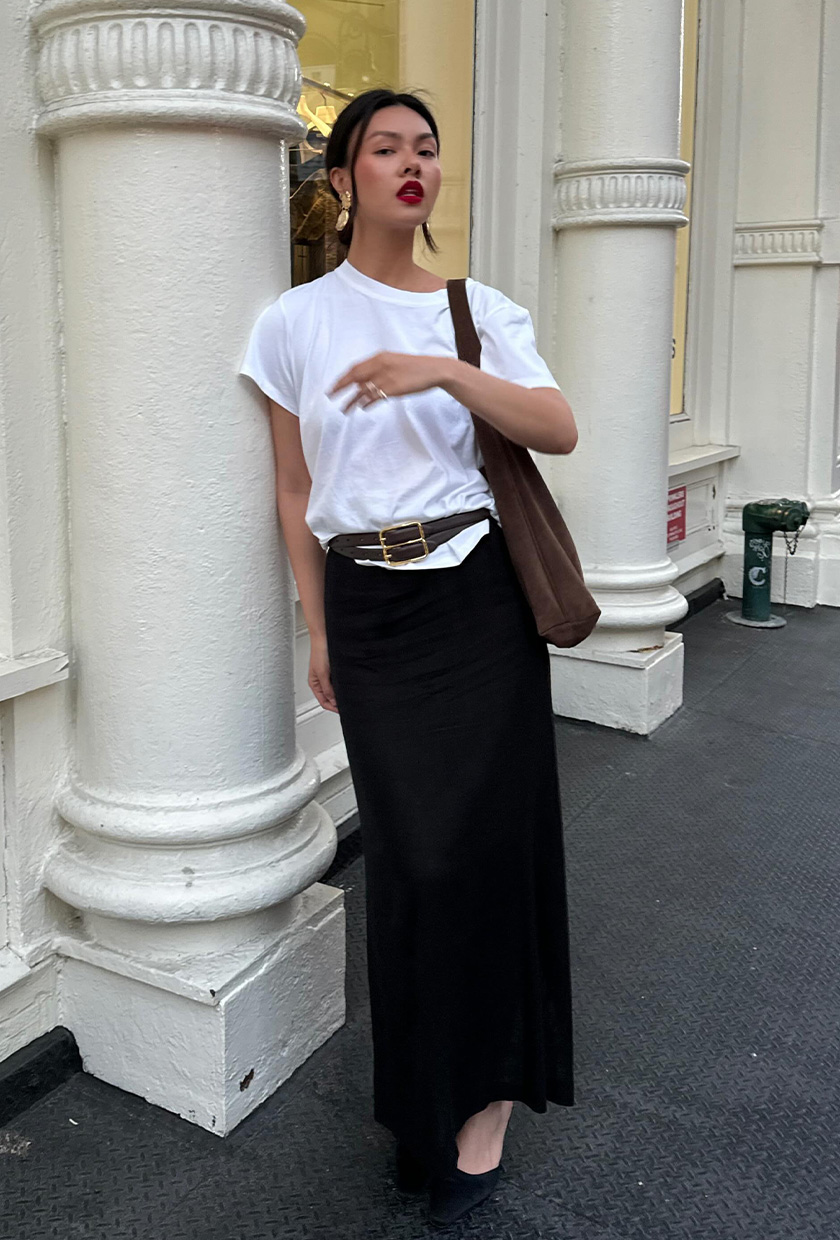 how to wear a statement belt is shown in a photo of a woman wearing a baggy white t-shirt with a black maxi skirt, black pumps, a brown suede hobo bag, and two brown belts layered on top of the shirt