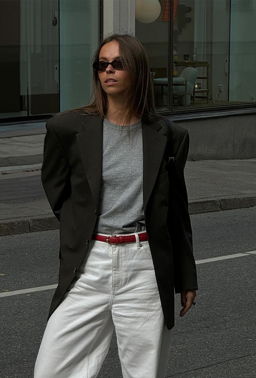 how to wear a statement belt is shown in a photo of a woman standing outside wearing a dark gray blazer over a light gray t-shirt with white jeans and a red statement belt