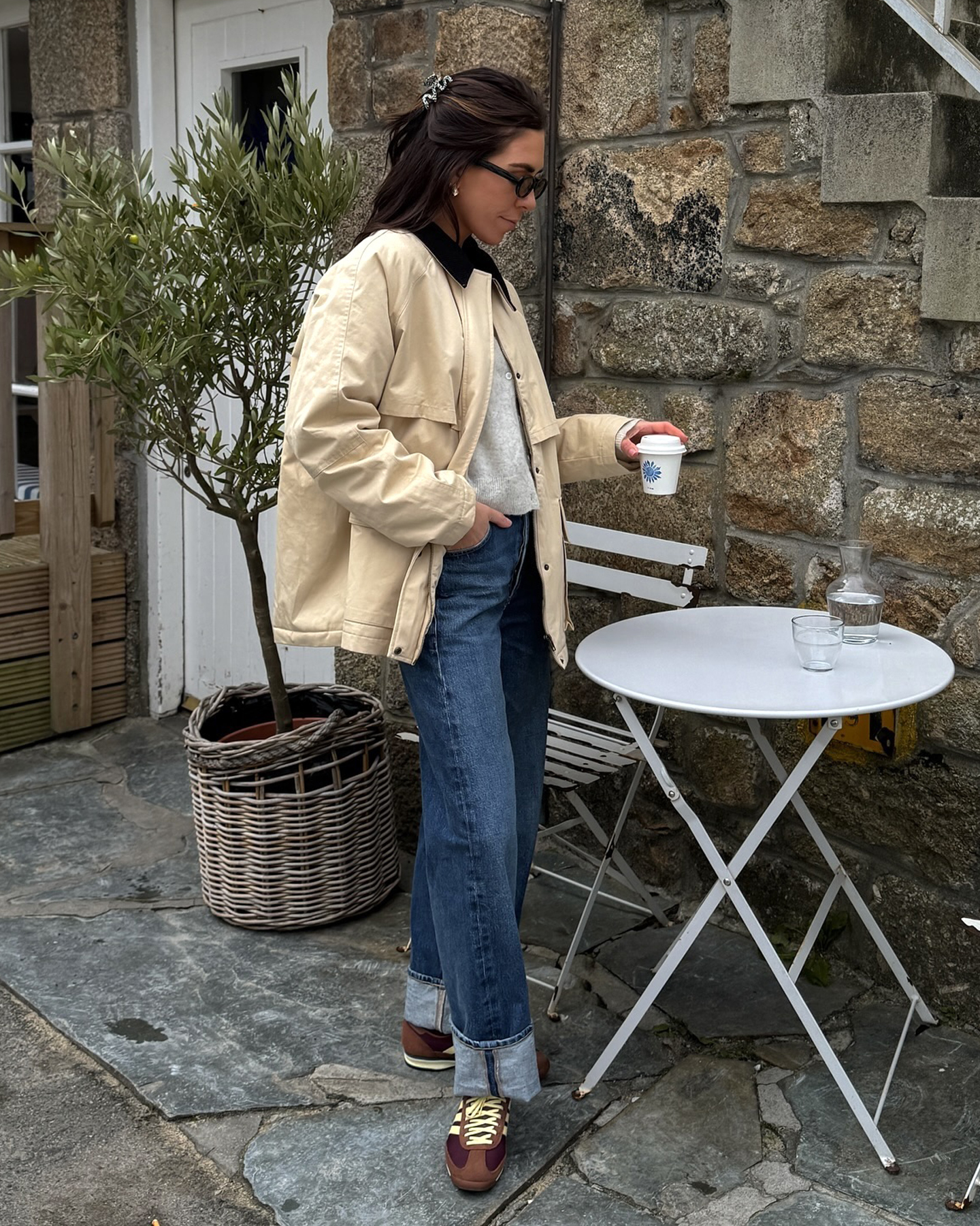 British influencer Jessica Skye poses at an outdoor cafe in Cornwall holding a mini coffee wearing a claw clip, sunglasses, a barn jacket, lightwieght cardigan, cuffed jeans, and brown Adidas SL72 sneakers