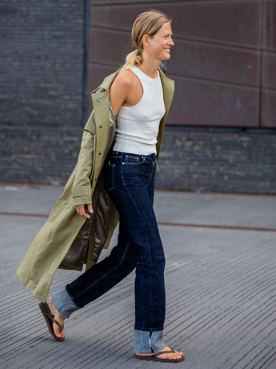 street style shot of Danish model Laura Julie during Copenhagen Fashion Week wearing a green trench coat, white racerback tank top, dark-wash cuffed jeans, and brown flip-flop sandals