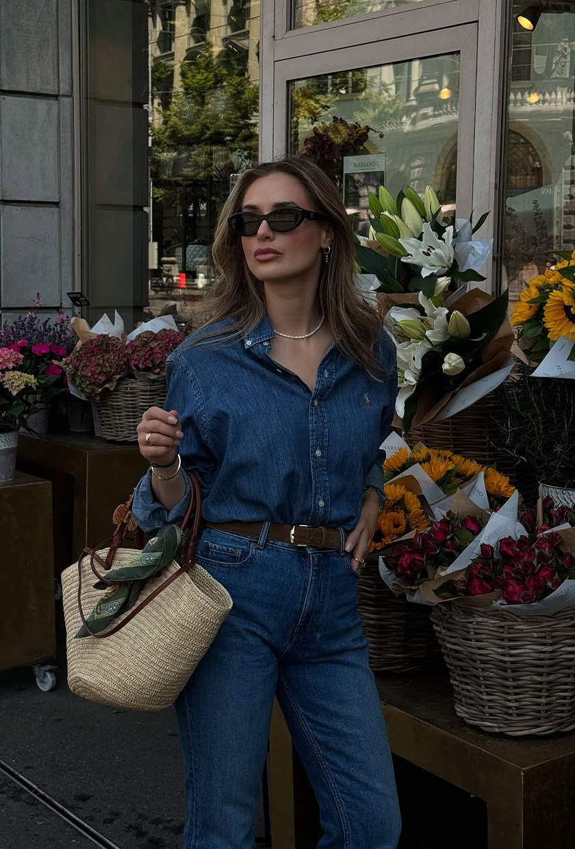 How to wear a statement belt is shown in a photo of a woman standing outside near a flower market wearing a denim button-down shirt with matching high-waisted jeans and a dark brown suede statement belt