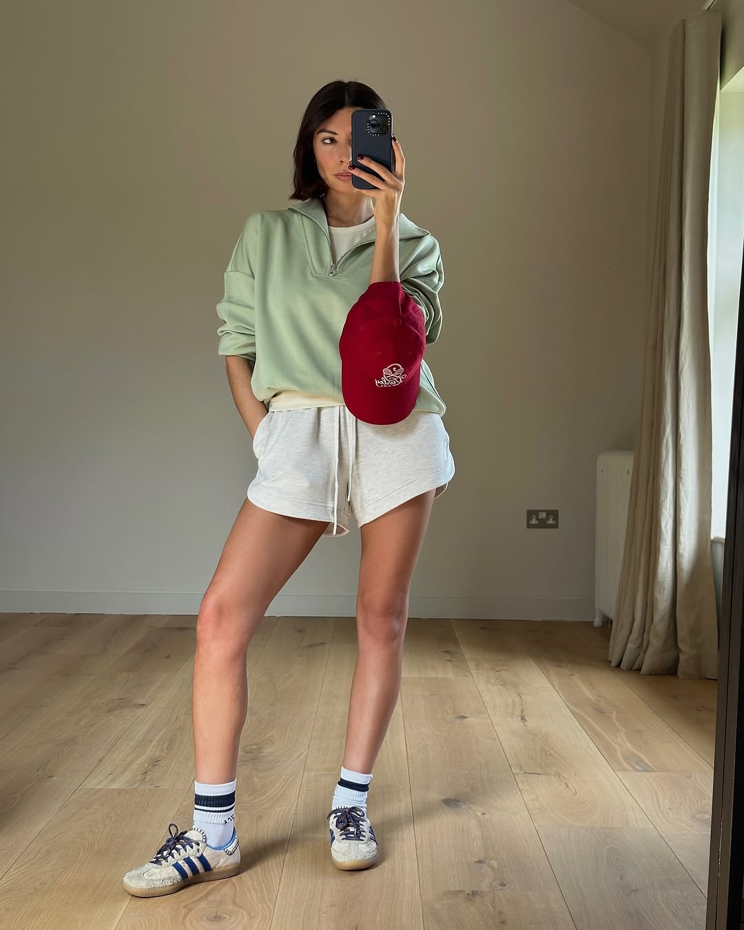 Woman wearing green pullover sweatshirt with shorts, Adidas sneakers, and red baseball hat in a summer 2025 outfit.