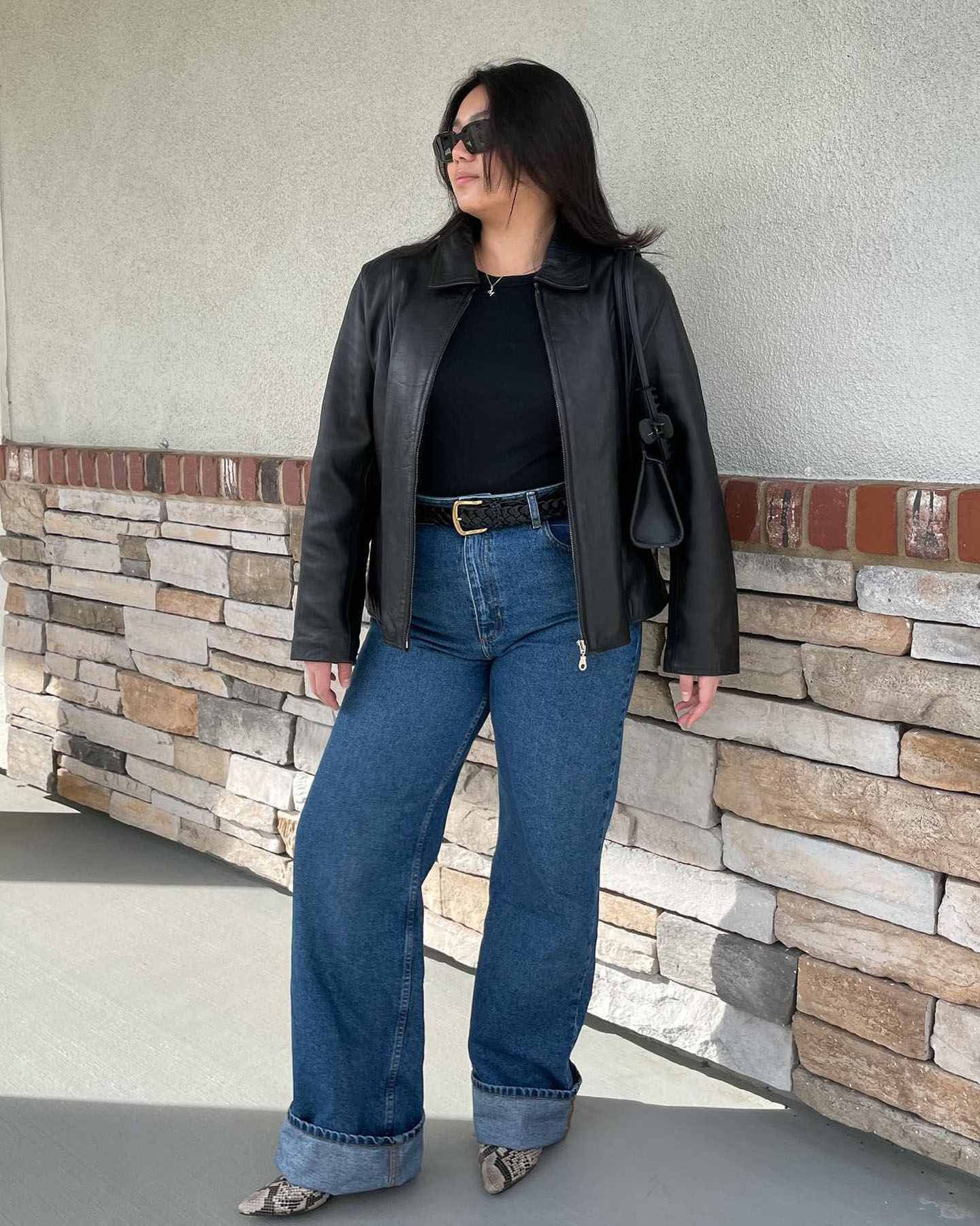 fashion influencer Marina Torres poses with a black leather jacket, black top, classic black belt, cuffed jeans, and snakeskin pointed-toe shoes