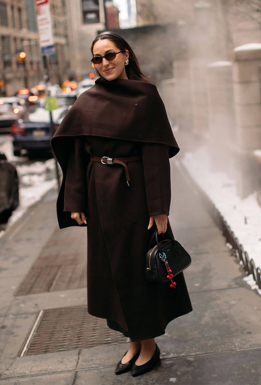 How to wear a statement belt is shown in a photo of a woman standing outside wearing a brown wrap coat with a brown statement belt styled on top paired with a black bowler bag, black ballet flats, a black headband, and black sunglasses