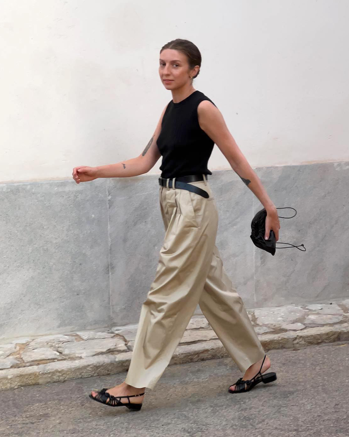 British influencer Brittany Bathgate walking on a street in Molico, Spain wearing a black crewneck tank top, black leather belt, minim black pouch clutch bag, pleated khaki pants, and black woven slingback sandals
