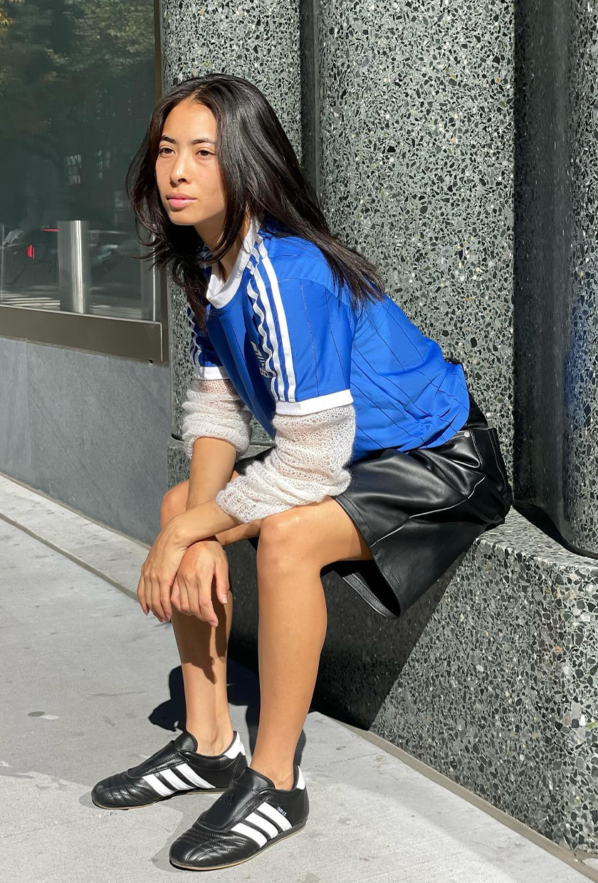 A woman wearing a blue short-sleeve sports jersey over a white long-sleeve gauzy knit shirt with leather Bermuda shorts and Adidas shoes.