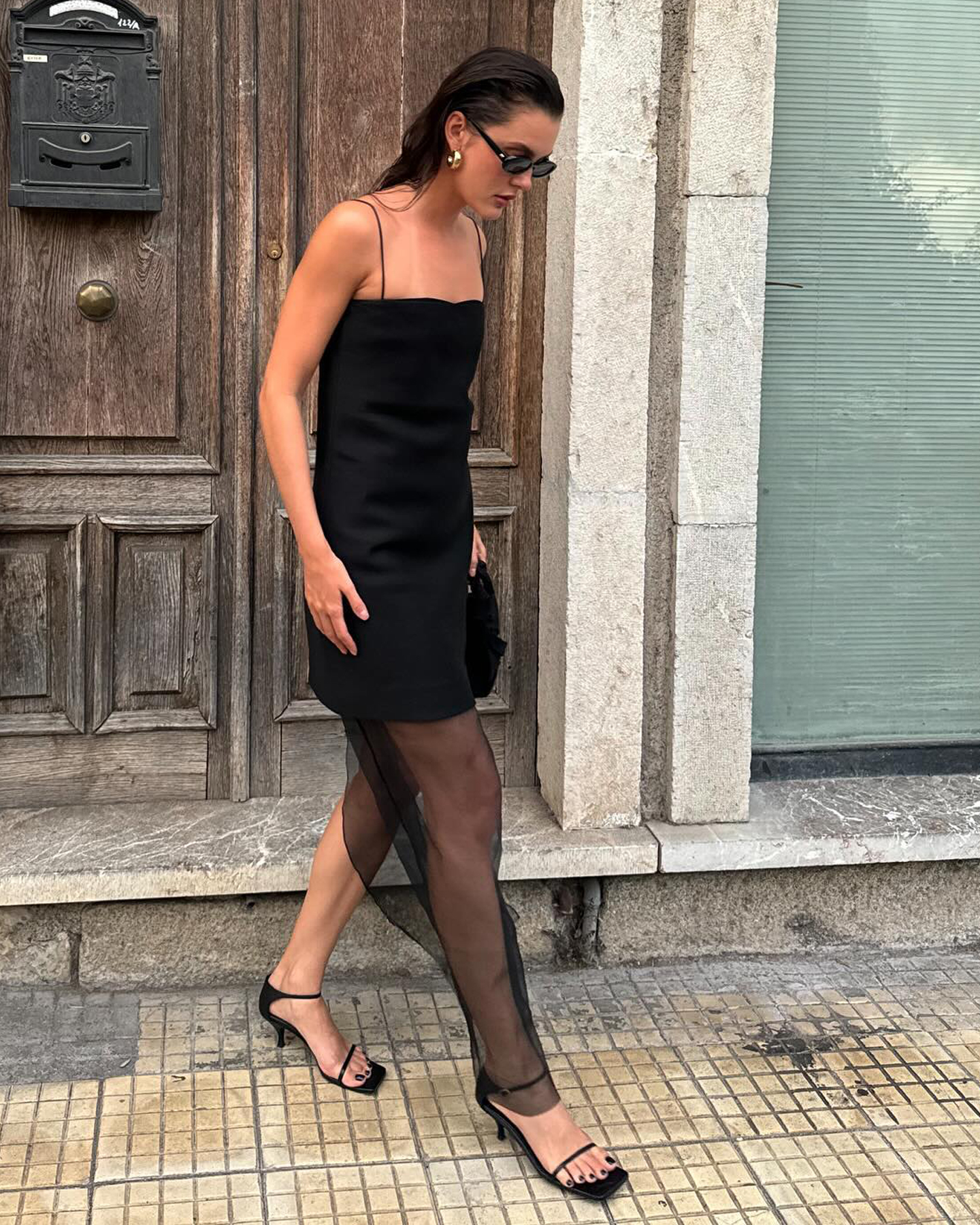 fashion influencer Valeria Soba wearing black sunglasses, a black cami mini dress with sheer hem, and black strappy minimalist heeled sandals.