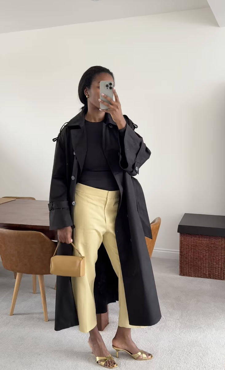 A woman wears a black trench, yellow trousers, and gold sandals.