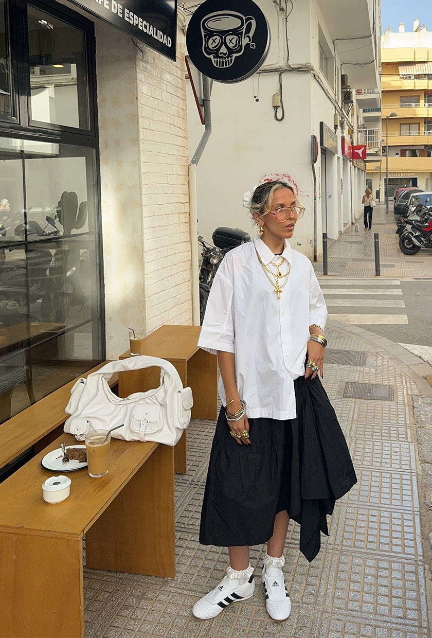 A woman standing outside a coffee shop and wearing an oversize white button-down shirt with gold layered chains, gold bangles, a black balloon skirt, white sneakers, and a white shoulder bag.