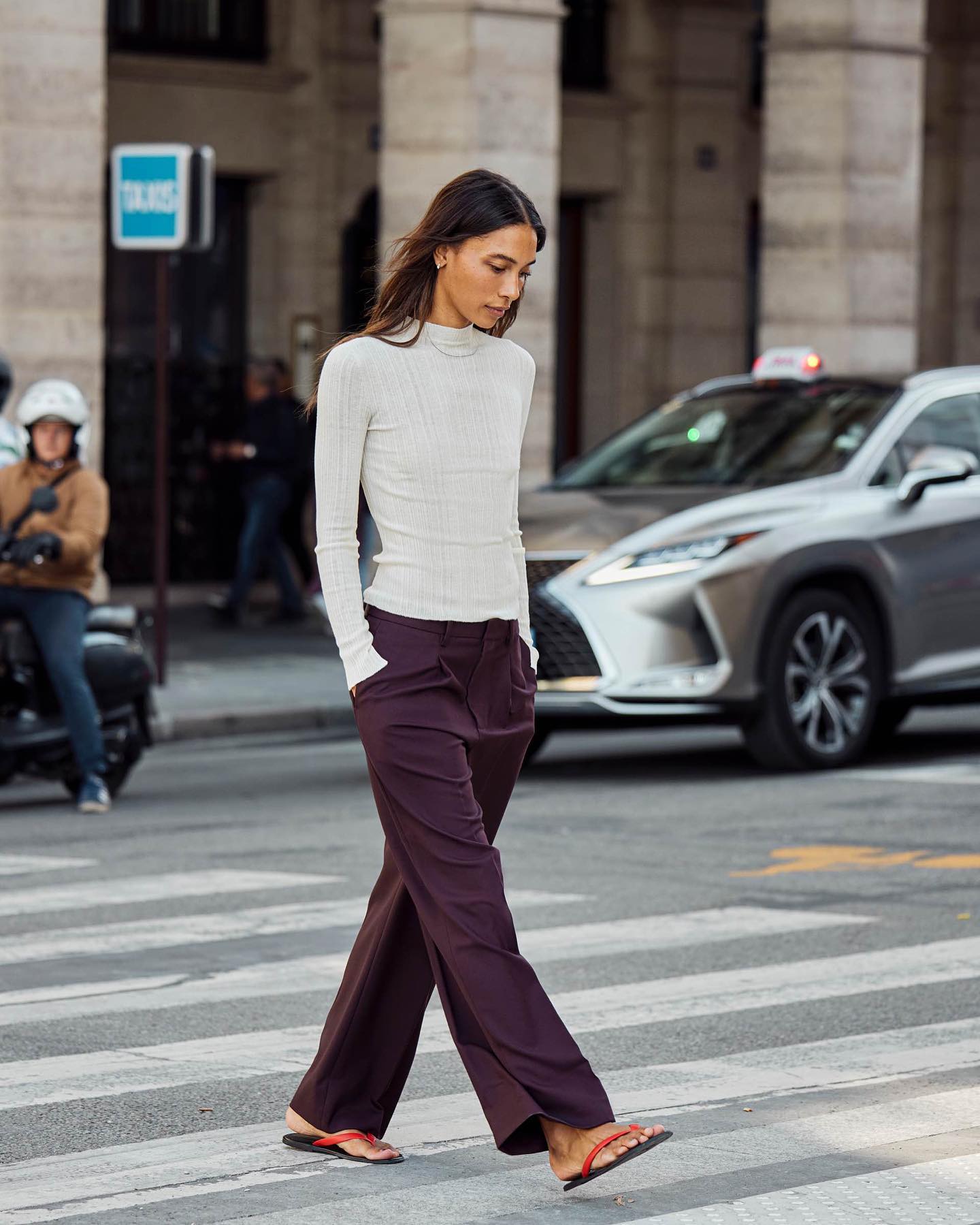 fashion influencer Tylynn Nguyen walking across a NYC crosswalk wearing a white ribbed long-sleeve top, eggplant colored trousers, and red and black flat flip-flop sandals.