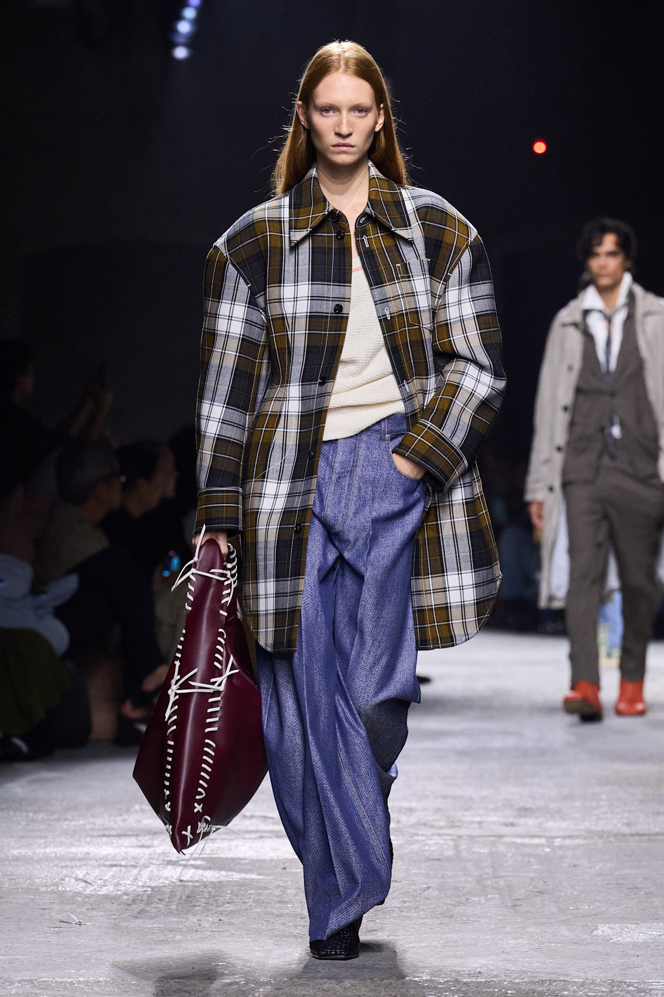A look from Bottega Veneta's S/S 25 Milan Fashion Week show including a plaid long-line jacket, blue jeans, and a burgundy bag. 