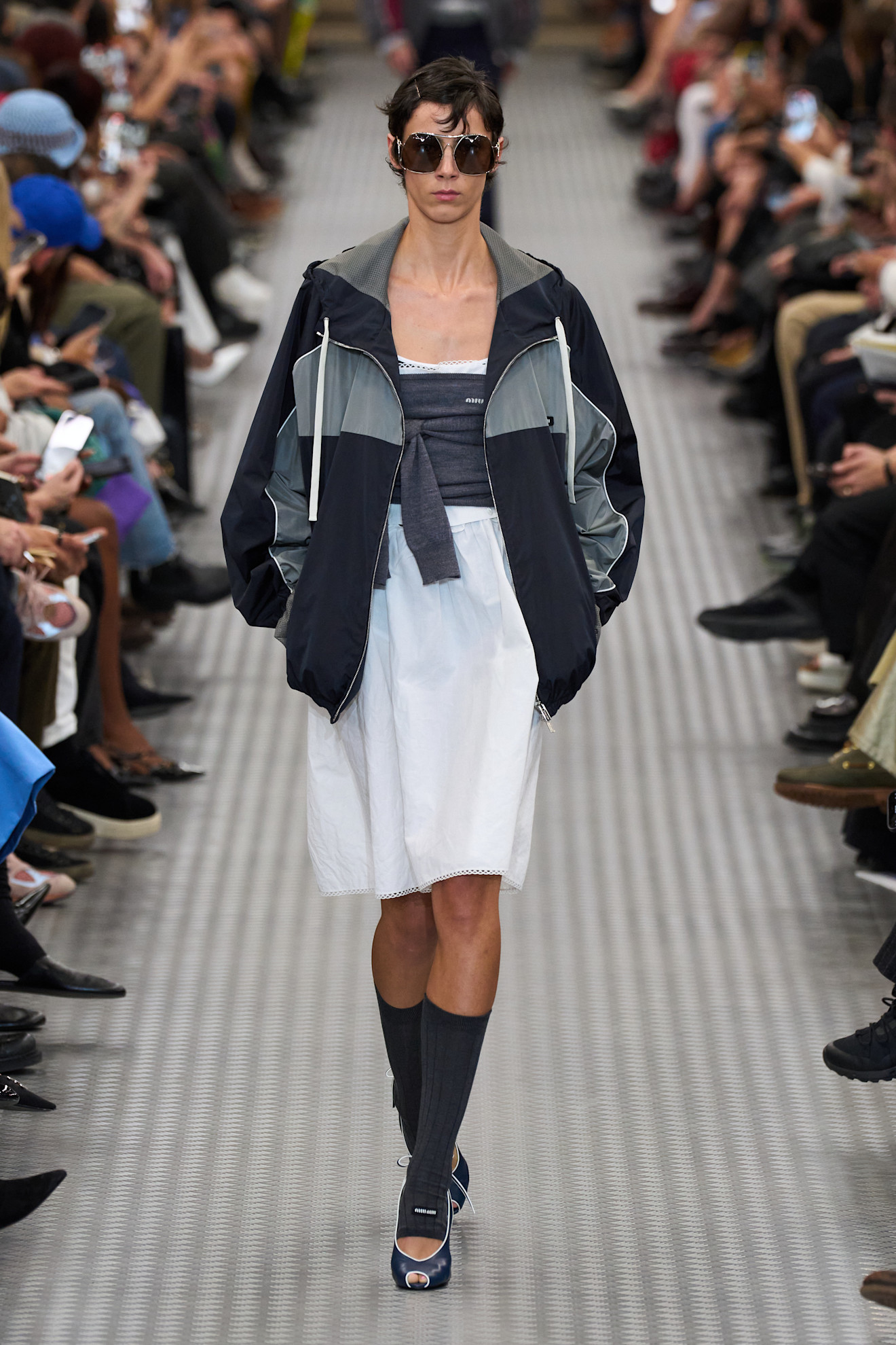 A S/S 25 Miu Miu model wearing a sporty zip-up jacket with a white poplin dress, socks, and peep-toe heels. 