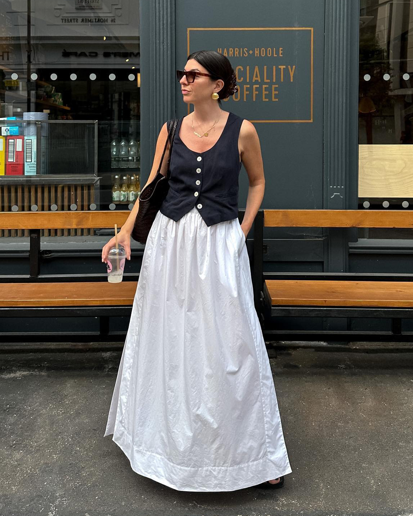 British style influencer Francesca Saffari poses outside of Harris + Hoole cafe in London wearing black sunglasses, statement earrings, button-front black vest, and a full white maxi skirt