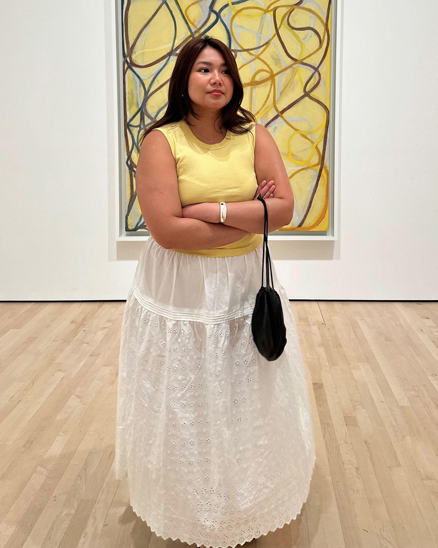influencer Marina Torres poses in front of an abstract yellow painting wearing a yellow tank top, silver bangle, black drawstring bag, and a white tiered eyelet maxi skirt
