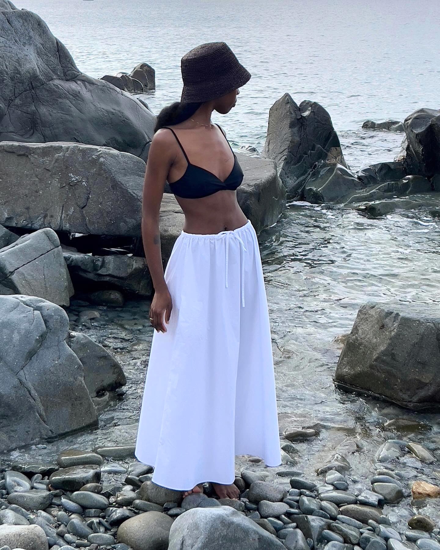 style influencer Sylvie Mus poses on rocks on on a beach in the South of France wearing a raffia black bucket har, black bikini top, and full white maxi skirt