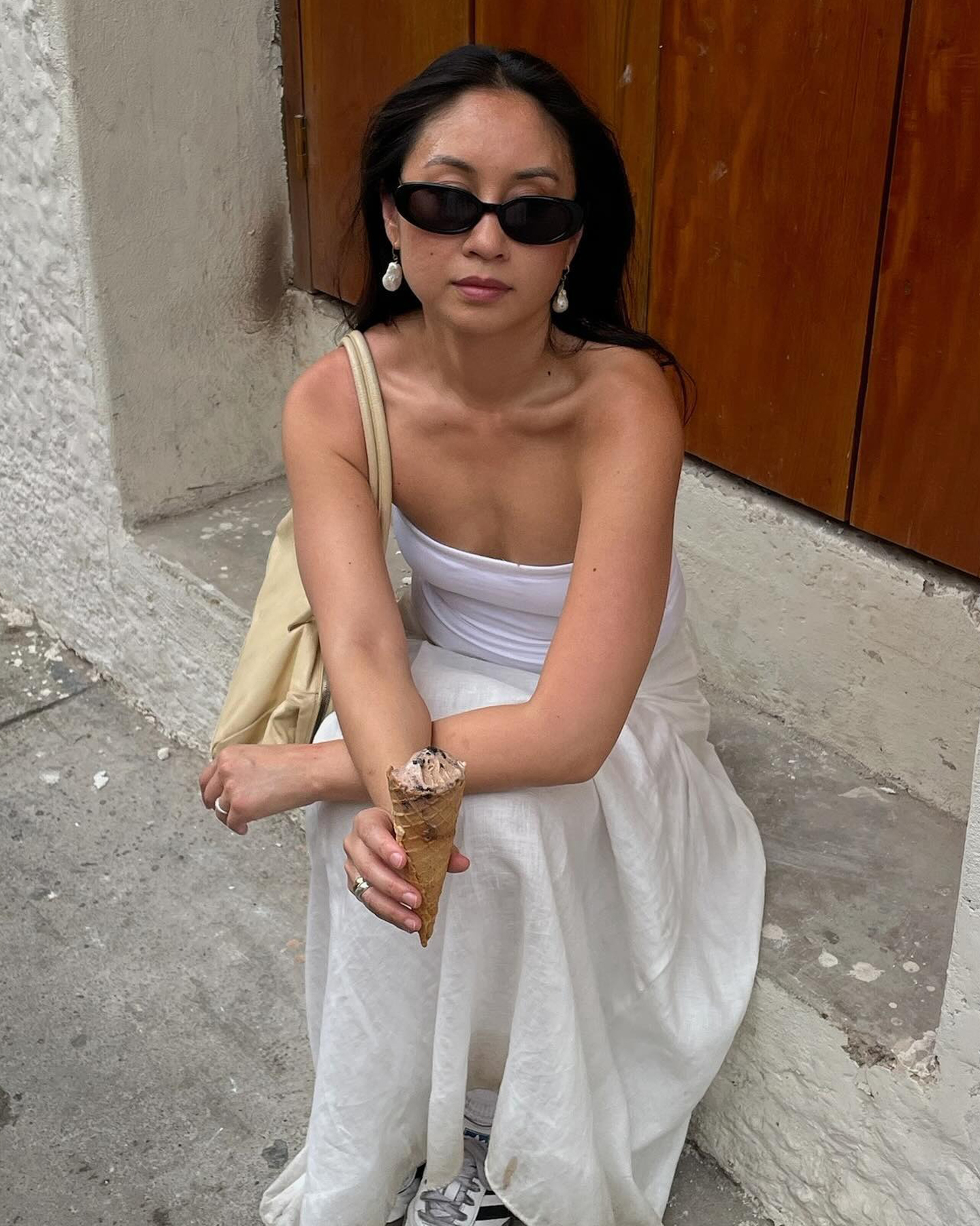 style influencer Jordan Risa sits on a step on a sidewalk in Cartagena, Colombia with black oval sunglasses, drop pearl earrings, a butter-yellow shoulder bag, white tube top, white linen skirt, and Adidas sneakers