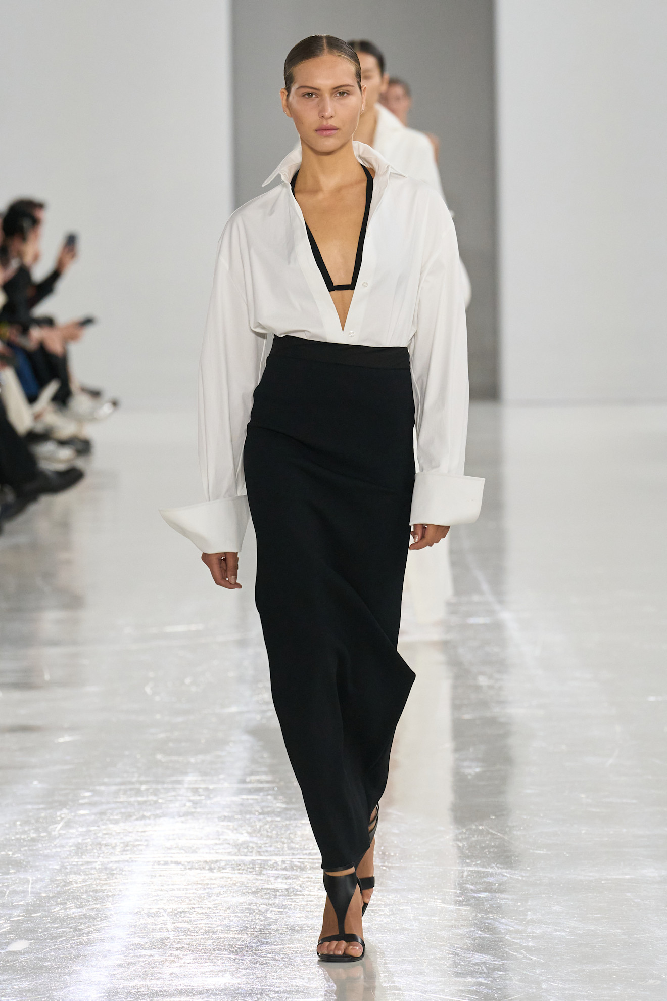A S/S 25 Max Mara model wearing a black bikini, white button-down shirt, black column skirt, and gladiator sandals. 