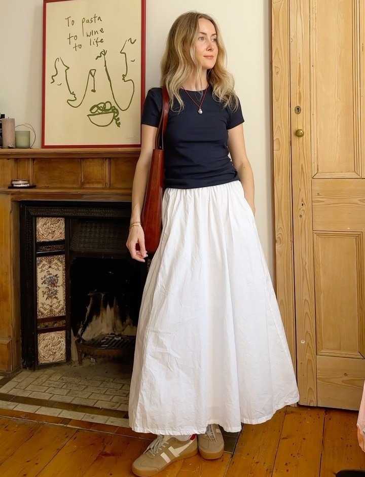 style influencer Chloe Butler poses in her London home wearing a cord pendant necklace, black short-sleeve tee, a red shoulder bag, full white skirt, and tan suede Gola sneakers