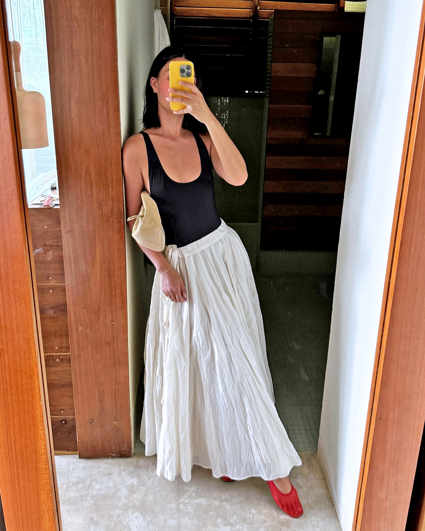 fashion influencer Mimi Nguyen poses for a mirror selfie while on vacation in Tulum Mexico with a bright yellow iphone case wearing a low-cut black tank top, tan suede bag as a clutch, full white maxi skirt, and red mesh flats