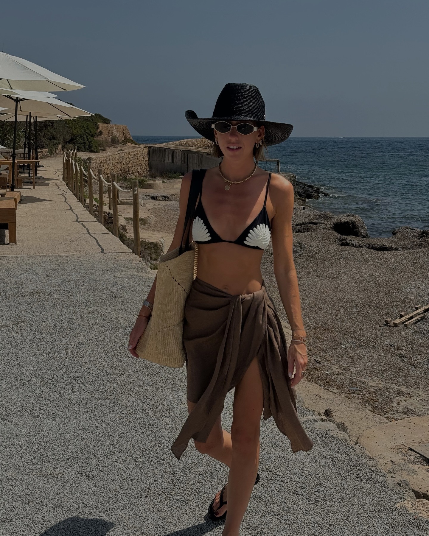 Hannah wears a black hat, black bikini, brown sarong and straw bag.