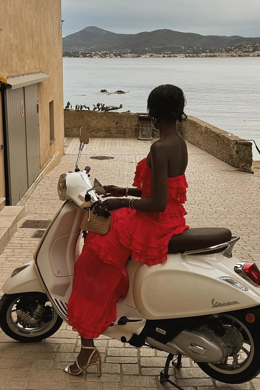 European vacation outfit idea is shown in a photo of a woman sitting on a moped wearing gold earrings, a strapless red ruffle maxi dress, a small raffia bag, and gold heeled sandals