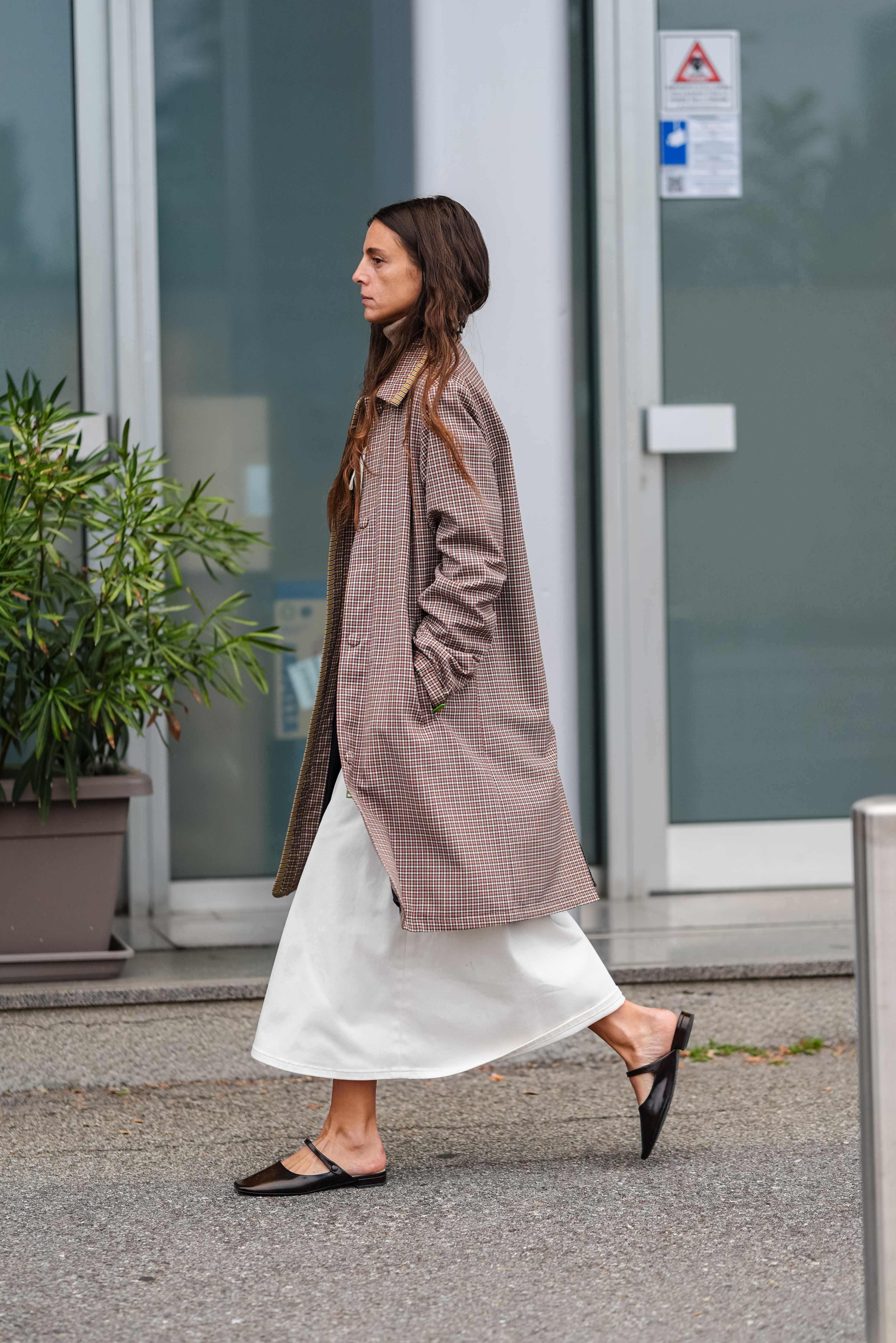 A woman wearing a pair of black leather mule flats, a white dress, and a brown plaid trench coat