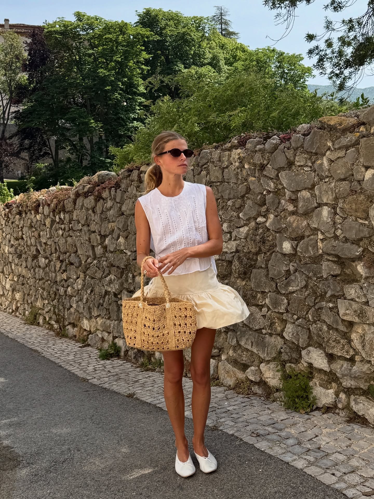 Alice wears a white top, yellow skirt, raffia bag, and white ballet flats.