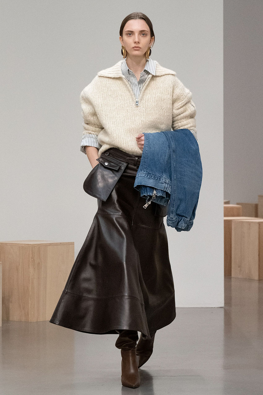 A Brandon Maxwell model wearing a knit cream sweater, brown leather skirt, and brown leather boots