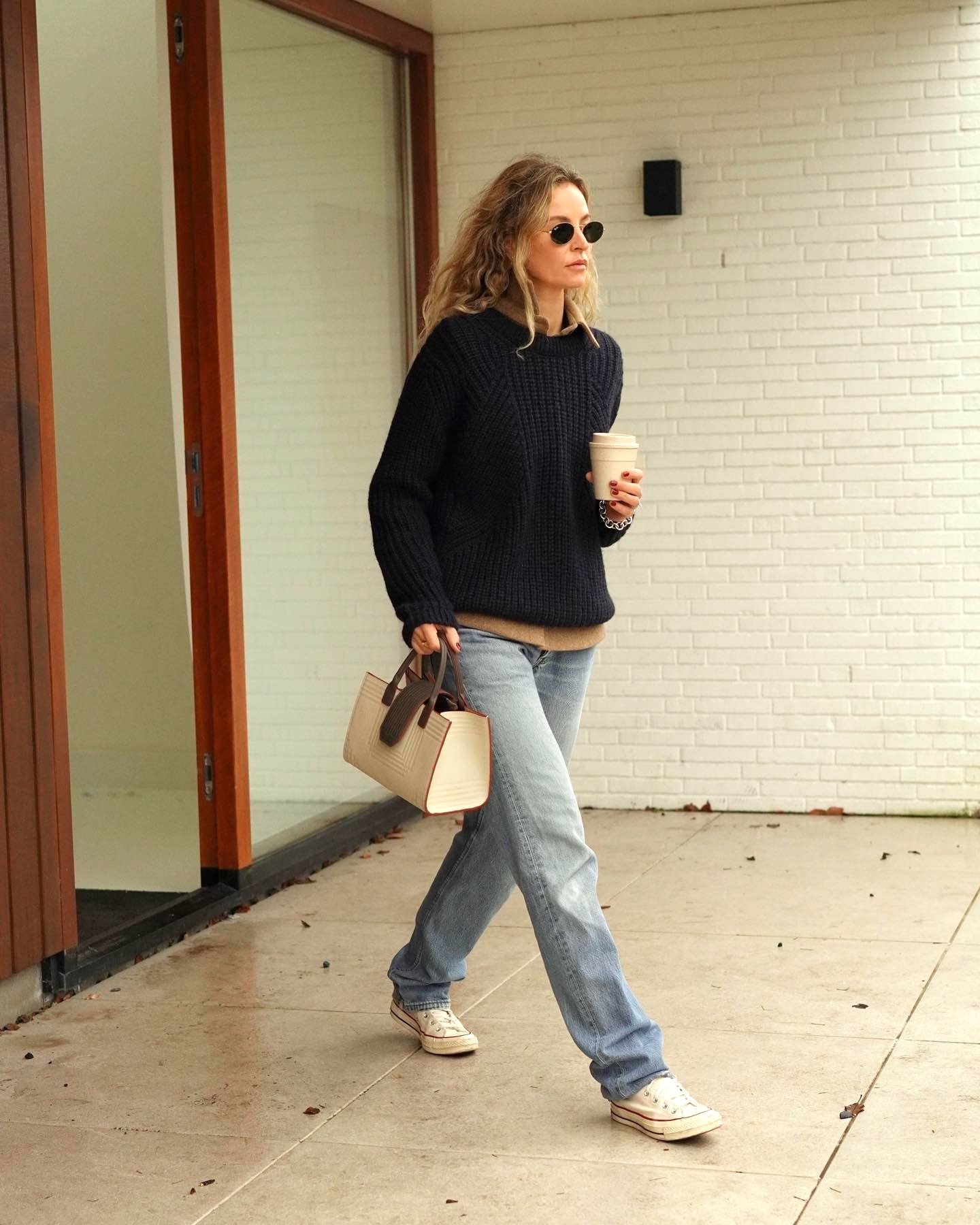 Style influencer Anouk Yve wearing round sunglasses, a layered ribbed sweater, a tan T-shirt, straight-leg jeans, and Converse sneakers.