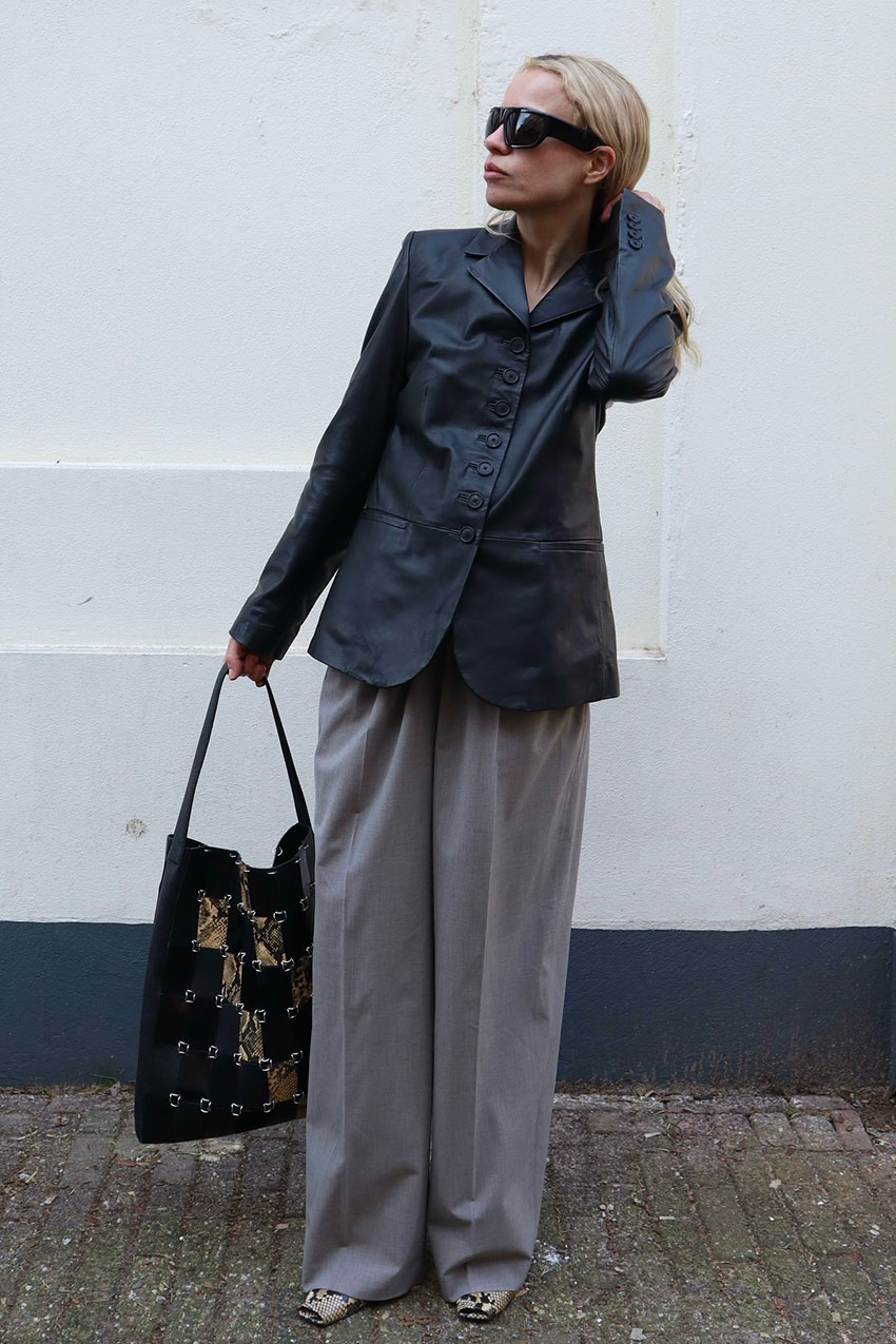 A woman wearing a black leather blazer, grey pants, a grommet black bag, and snakeskin boots
