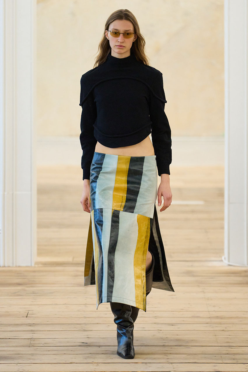 An Eckhaus Latta model wearing a black long sleeve top, striped skirt, and leather black boots