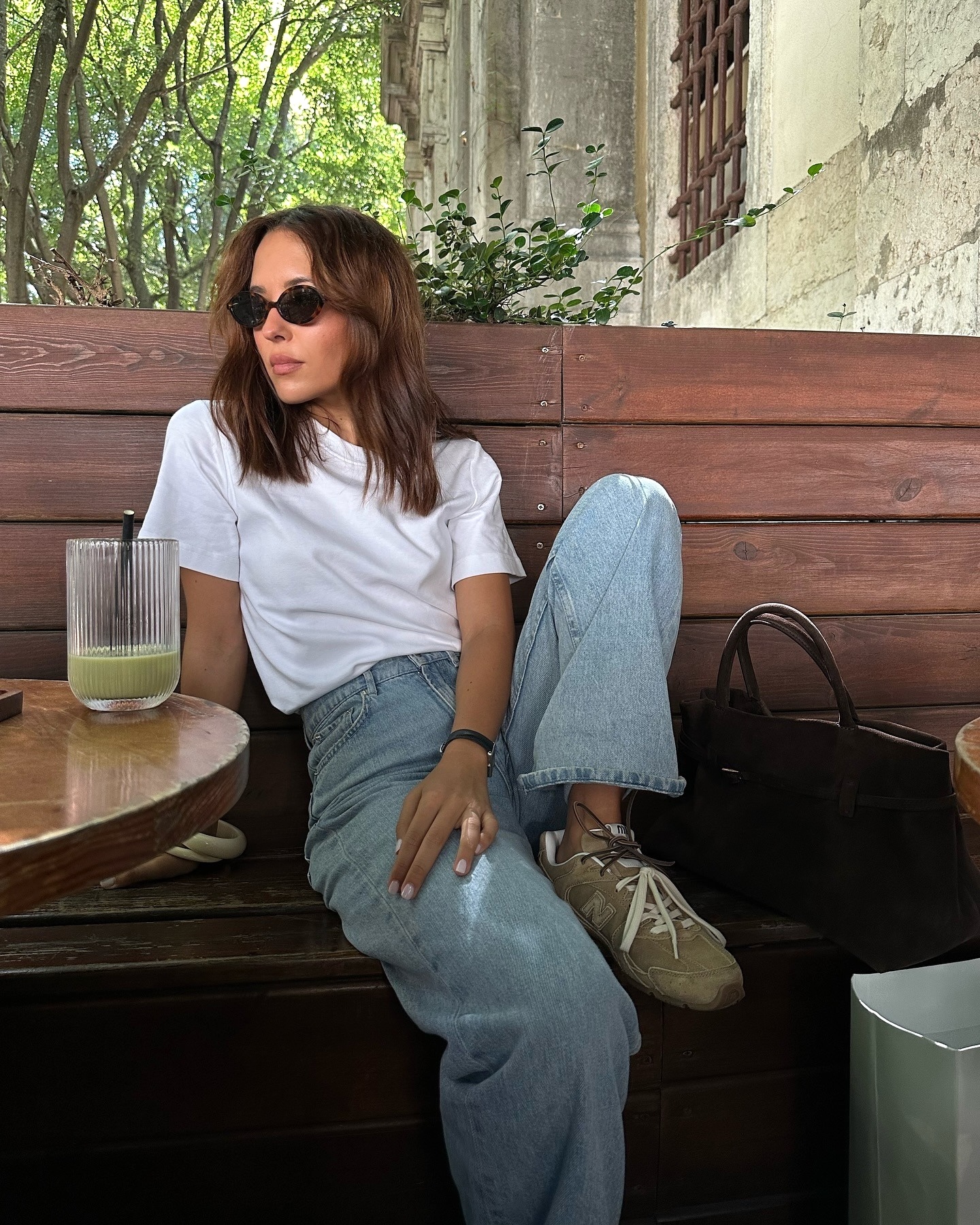 Fashion influencer Debora Rosa leaning on a bench in an outdoor cafe wearing oval sunglasses, a white T-shirt, straight-leg jeans, and New Balance x Miu Miu suede sneakers.