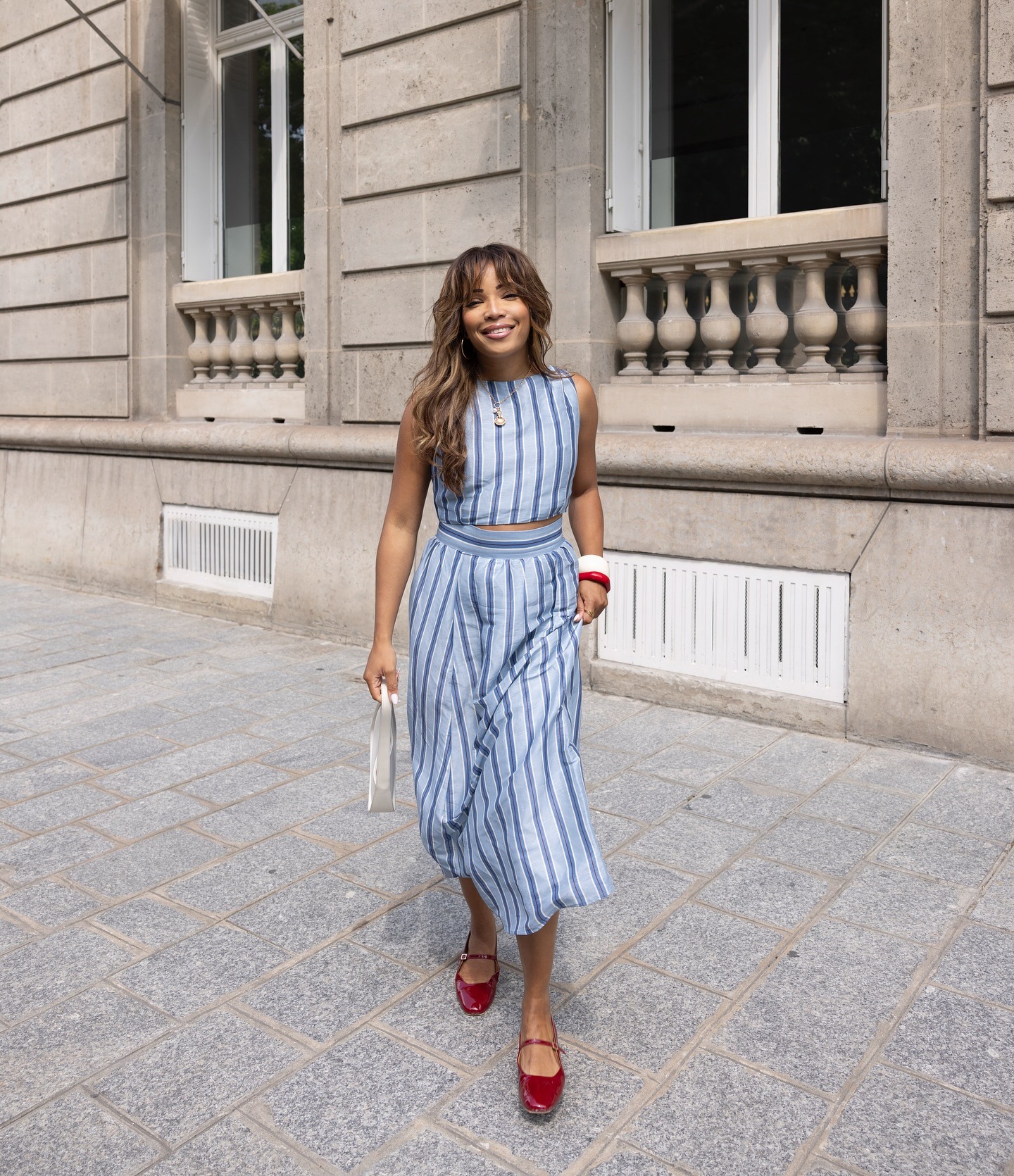 A woman wears a blue stripe top and skirt set with red mary janes.