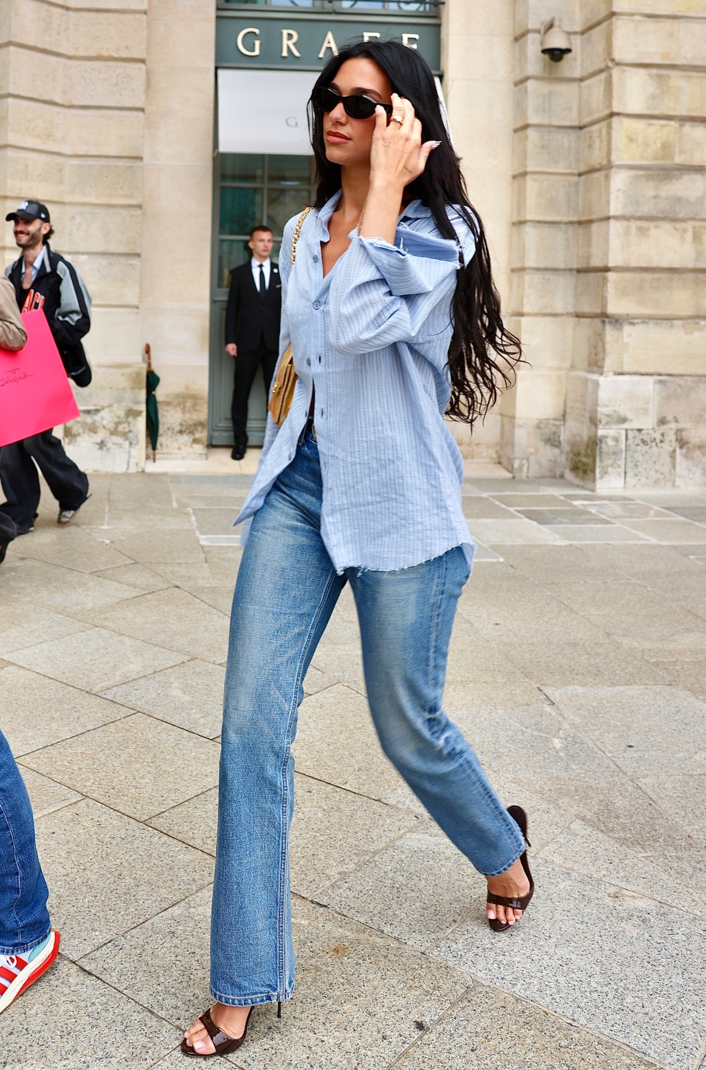 Dua Lipa in Paris wears a pair of straight-leg light-wash jeans with a blue button-down and open-toed mules.